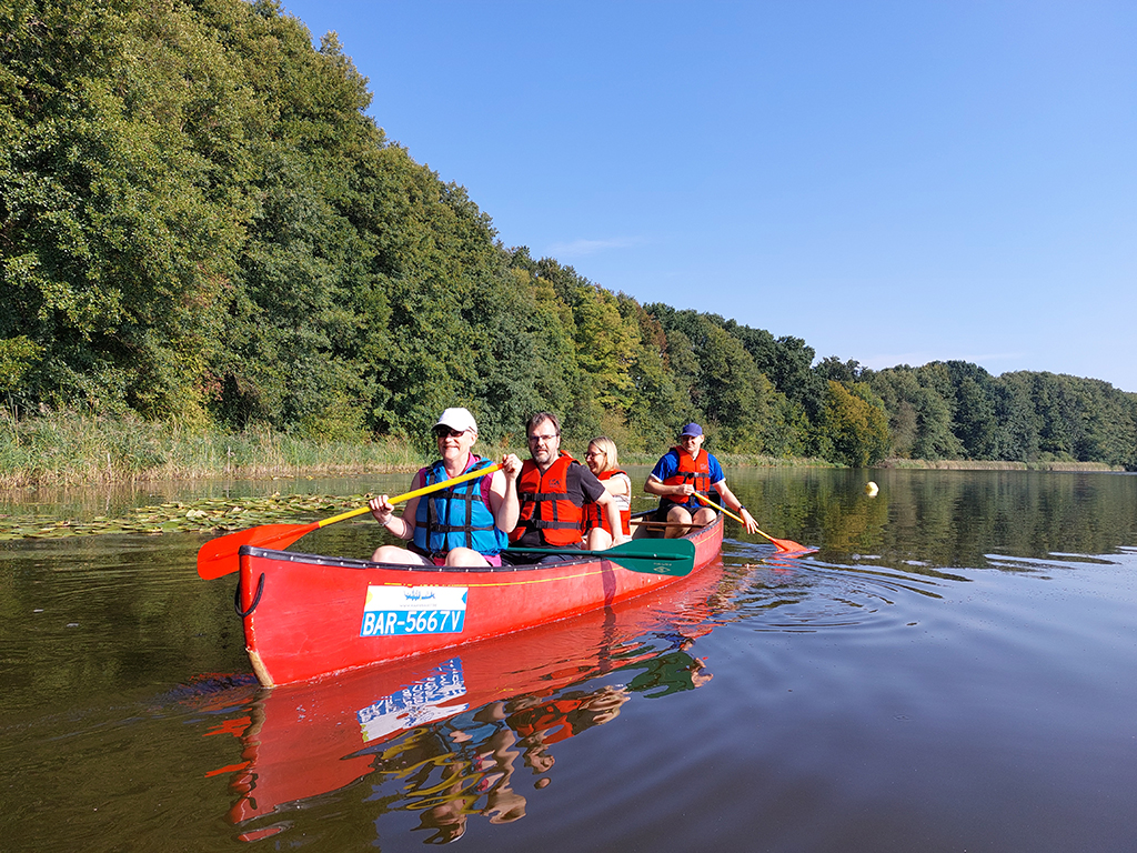 Teammitglieder paddeln gemeinsam in einem roten Kanu auf einem ruhigen See, umgeben von grünen Bäumen. Das Bild strahlt Zusammenhalt und Freude an der Natur aus.