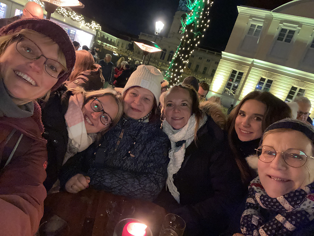 Ein fröhliches Team auf einem Weihnachtsmarkt, das die festliche Atmosphäre bei einem gemeinsamen Selfie genießt. Alle tragen Winterkleidung und lächeln in die Kamera.