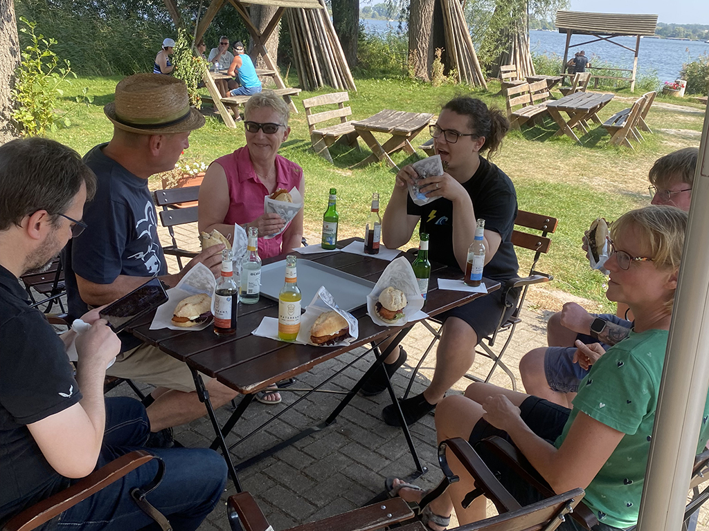 Teammitglieder sitzen an einem Tisch im Freien und genießen gemeinsam ihre Mahlzeit in entspannter, grüner Umgebung.