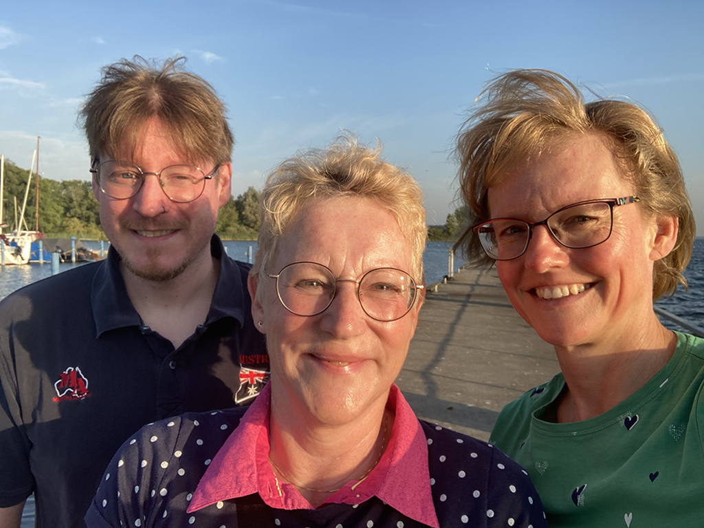 Drei Kolleg:innen machen ein Selfie am Pier, die Abendsonne beleuchtet ihre Gesichter und sorgt für eine warme Atmosphäre.